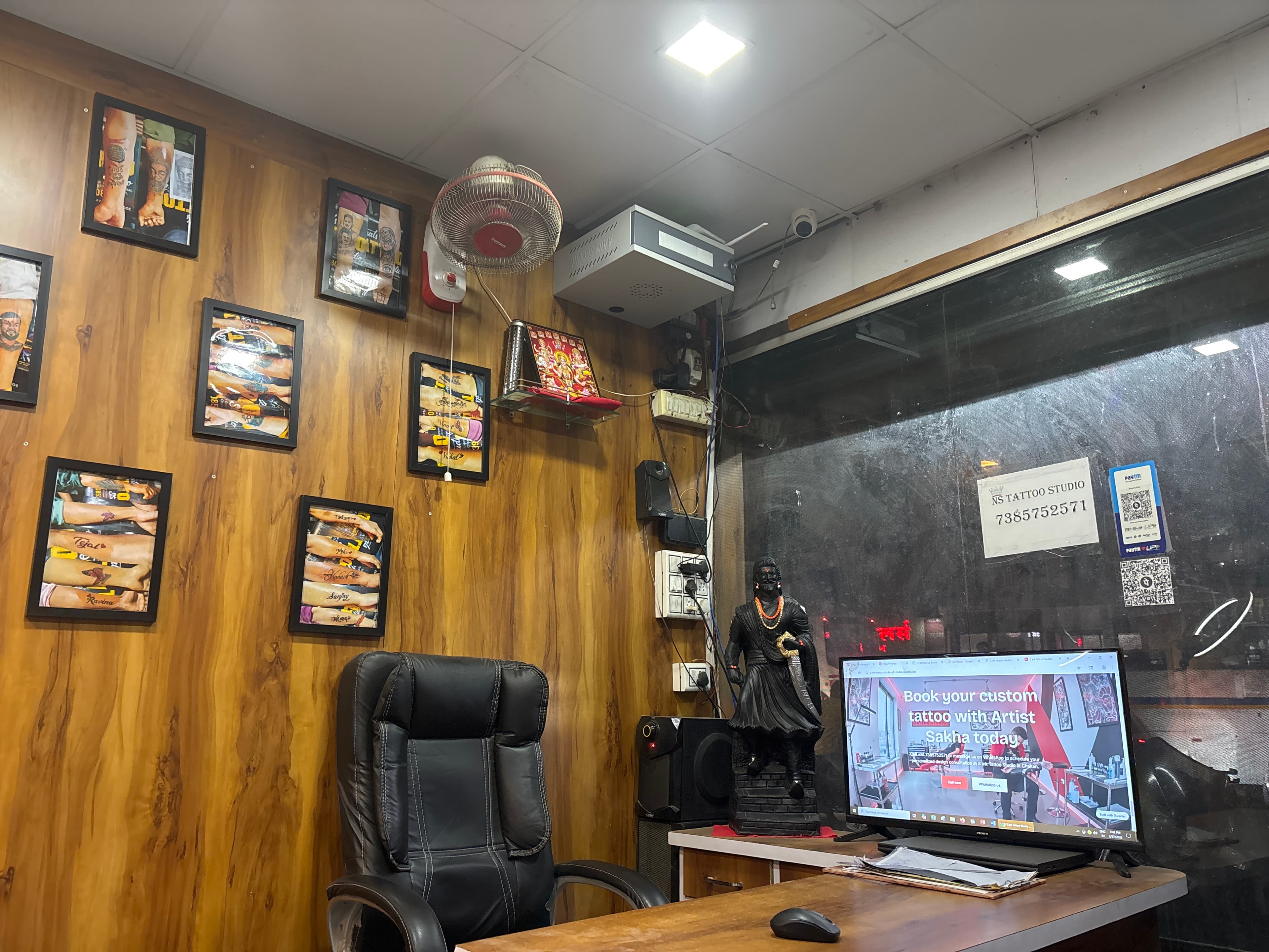 Wood-paneled tattoo studio interior with framed tattoo photos, a black chair, and a computer desk.